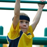 Child wearing a protective headband hanging from monkey bars during outdoor play.